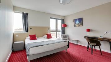 A hotel room with a bed, a desk, a chair, a lamp, and a painting on the wall.