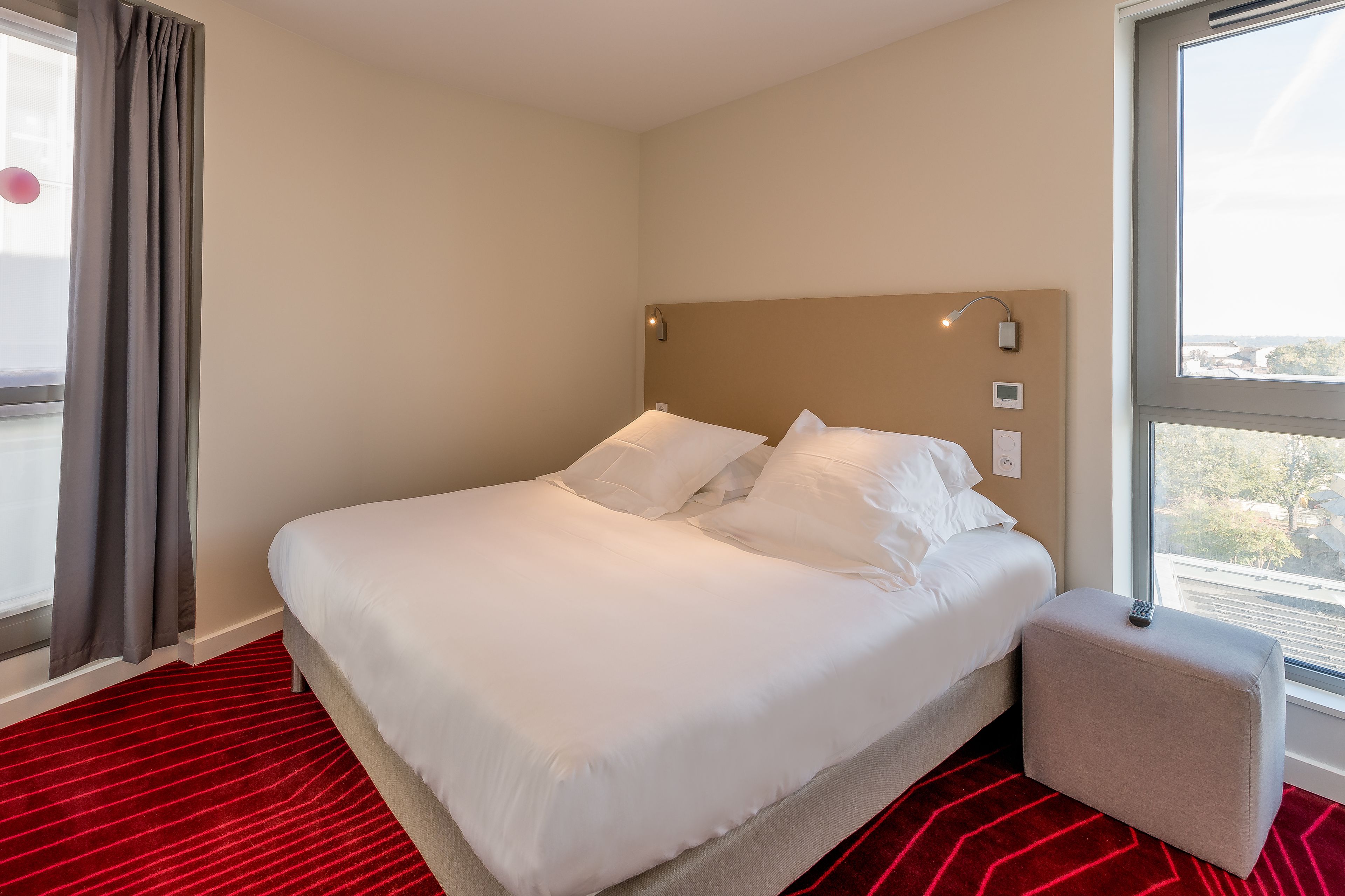 A hotel room with a bed, a red carpet, a window, and a view of the city.