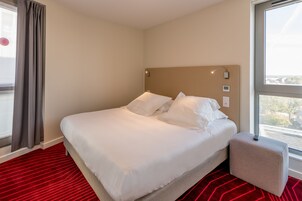 Camera d'albergo con un letto, un tappeto rosso, una finestra e vista sulla città.