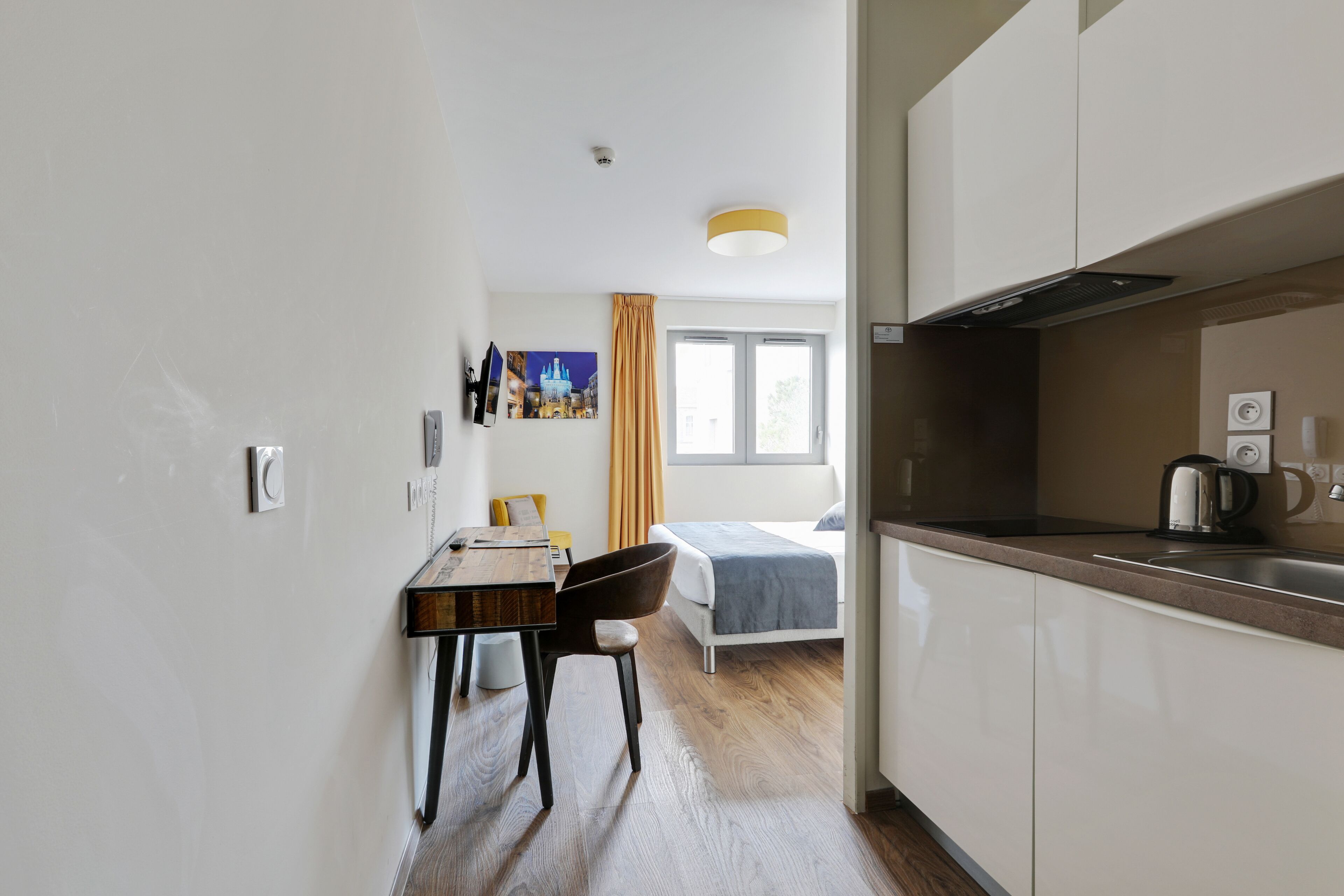 A modern hotel room with a wooden dining table, a single bed, and a kitchenette.