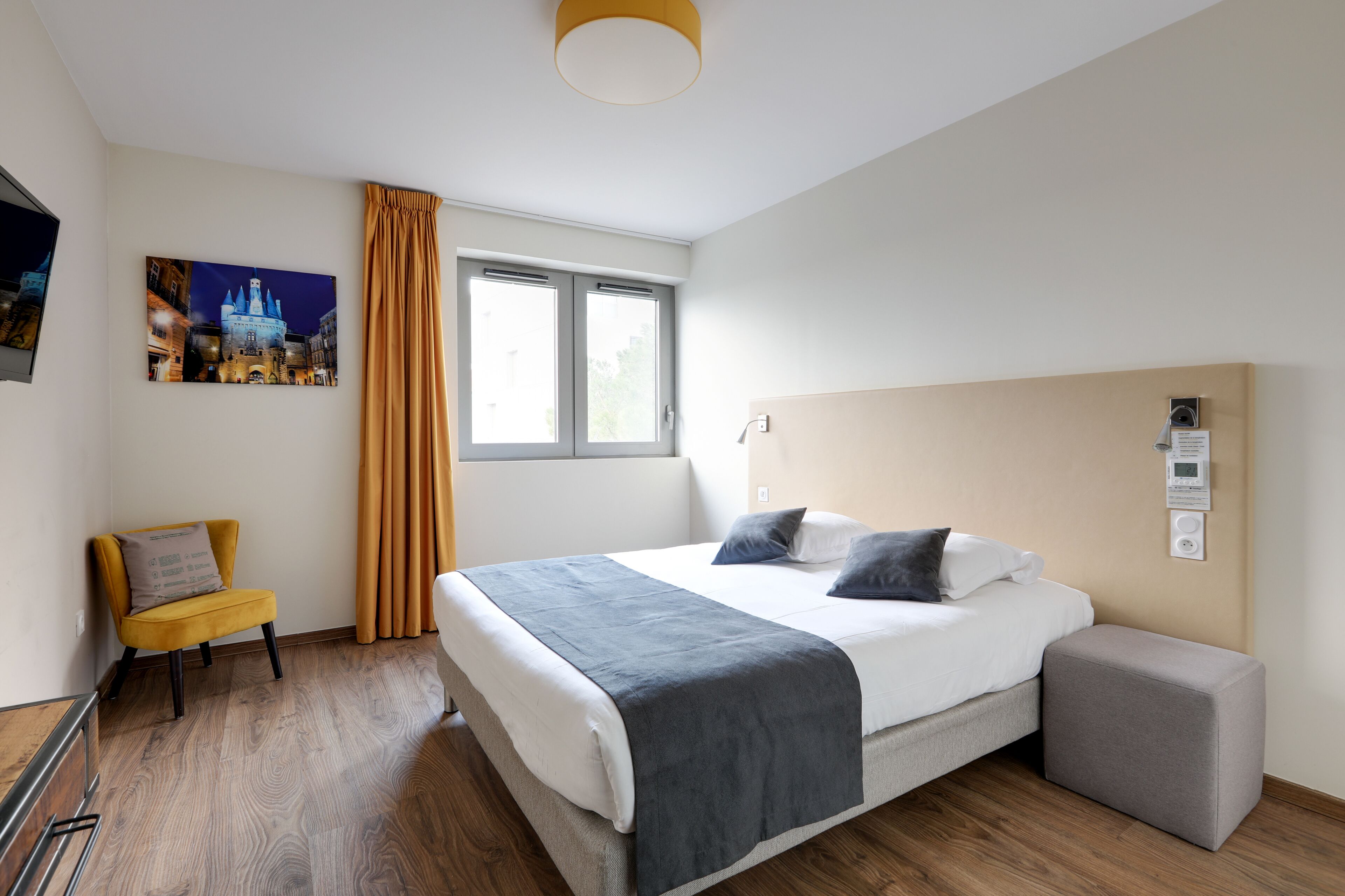 Una habitación de hotel moderna con una cama grande, un pequeño rincón de estar, una ventana con cortinas y una obra de arte colgada en la pared.
