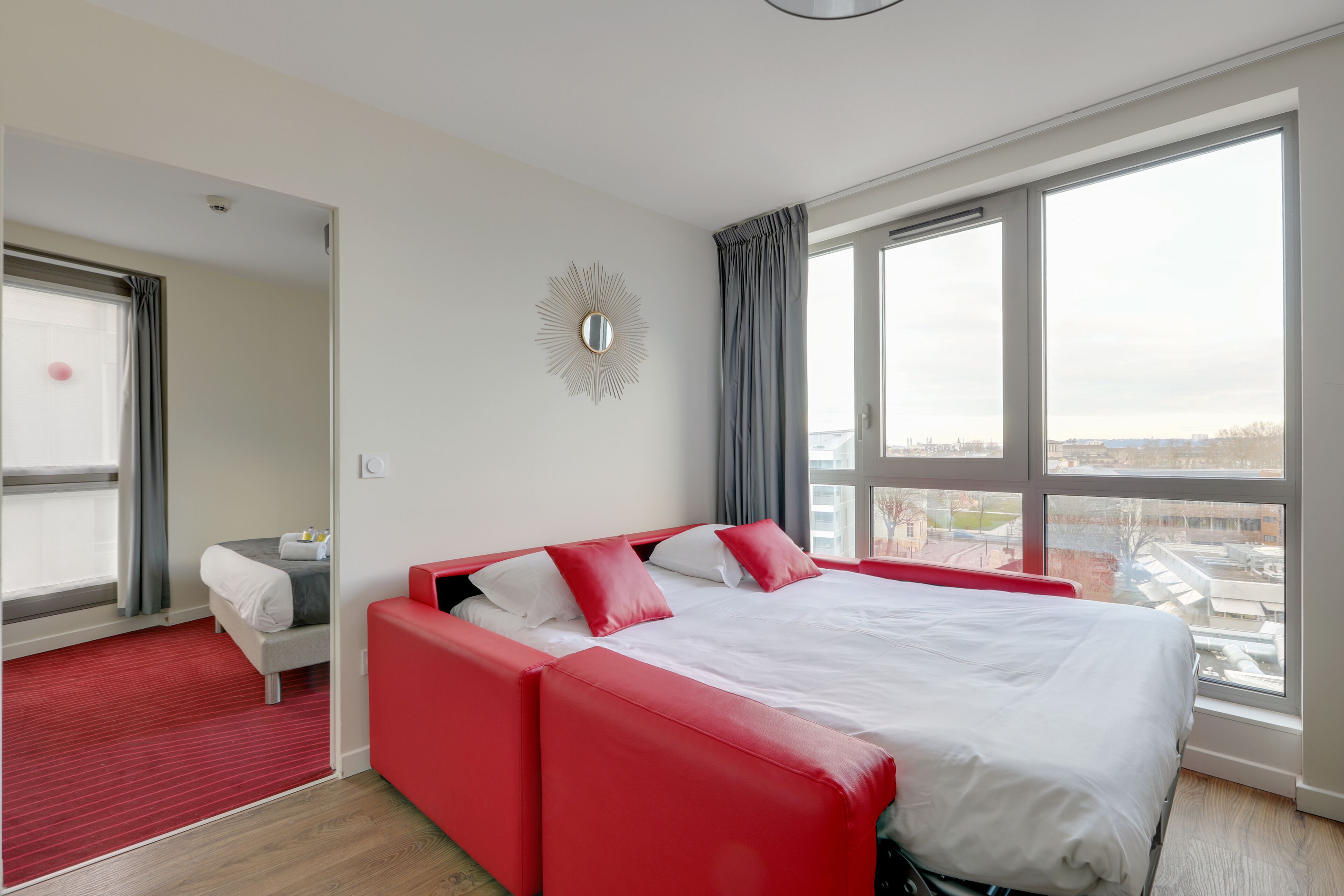 Une chambre d’hôtel moderne avec un grand lit, un canapé rouge, un petit bureau et une grande fenêtre donnant sur la ville.