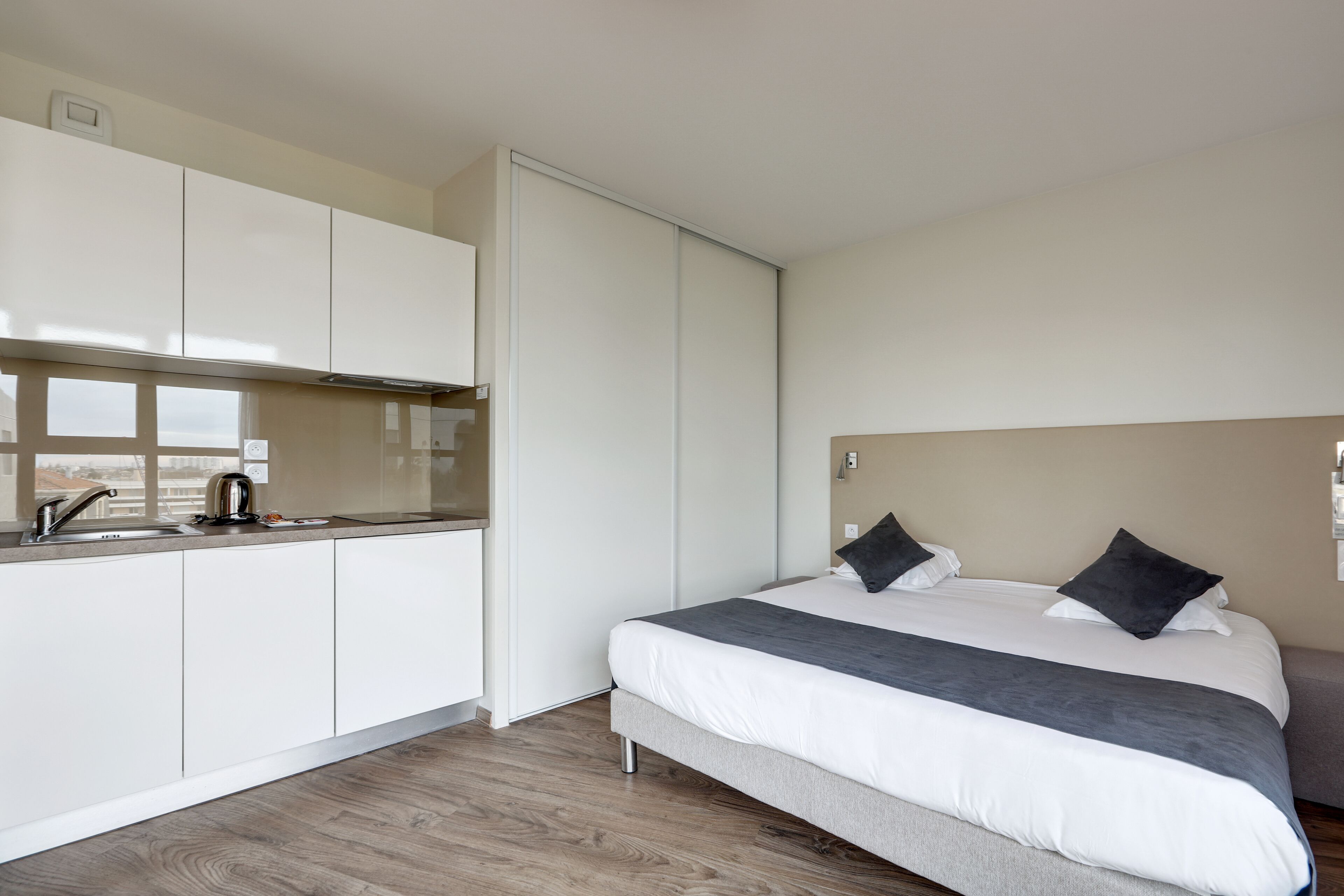 A modern hotel room with a kitchenette, a bed with two pillows, and a grey headboard.