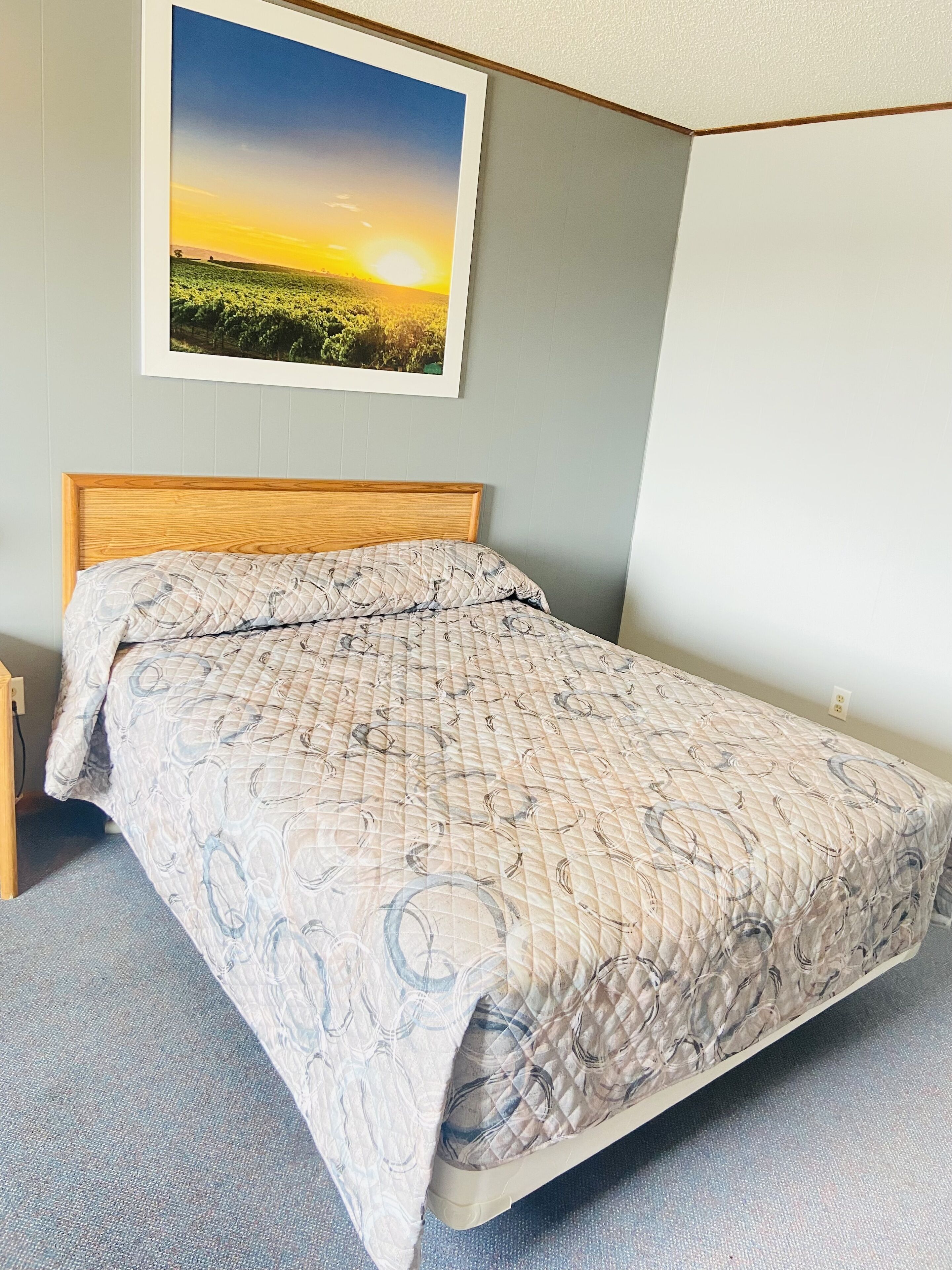Un lit bien fait, avec une couvre-lit à motifs, une tête de lit en bois et un tableau encadré représentant un coucher de soleil accroché au mur.