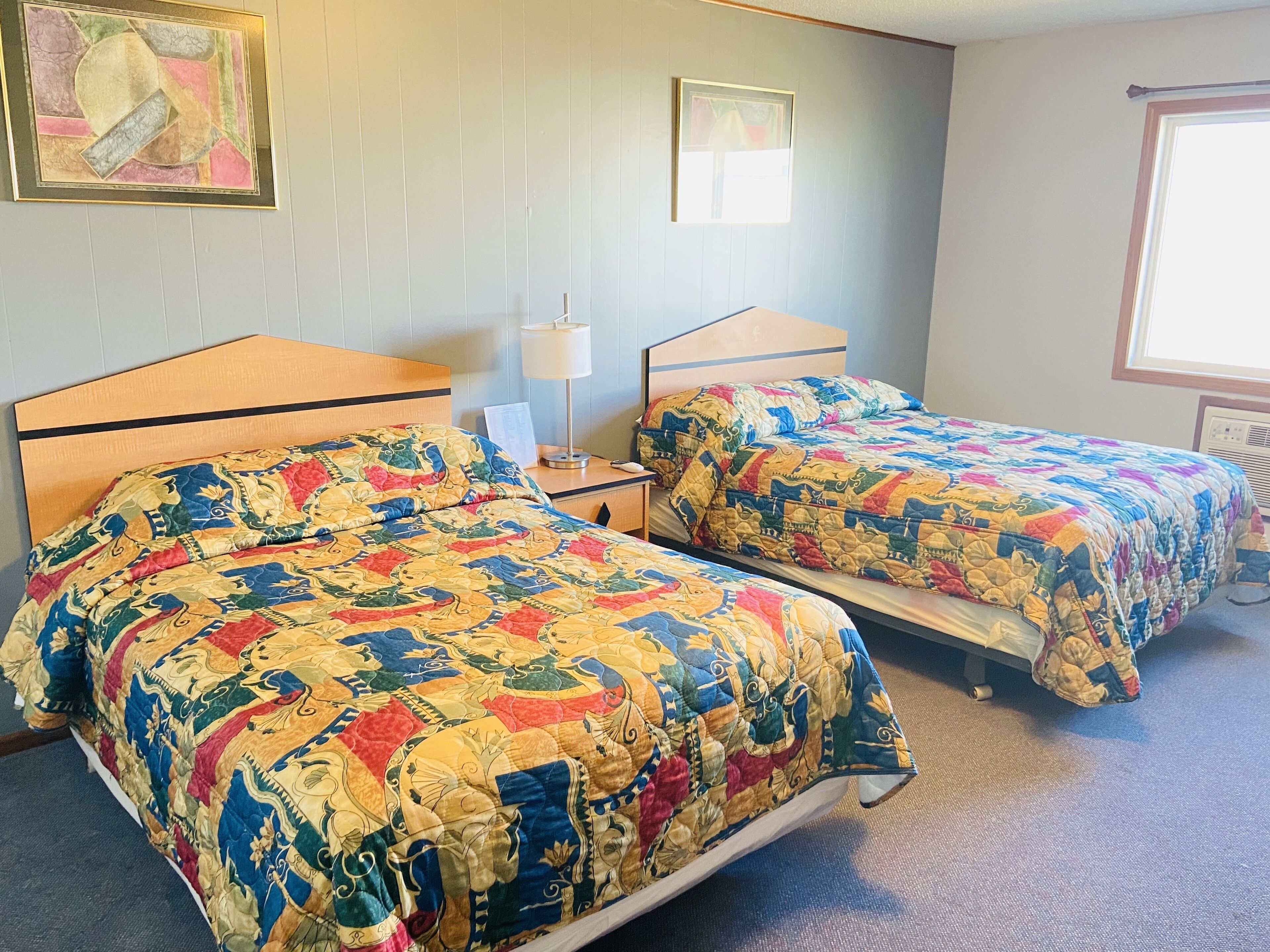 A hotel room with two beds, each with a colorful quilt, a bedside table with a lamp, and a framed picture on the wall.