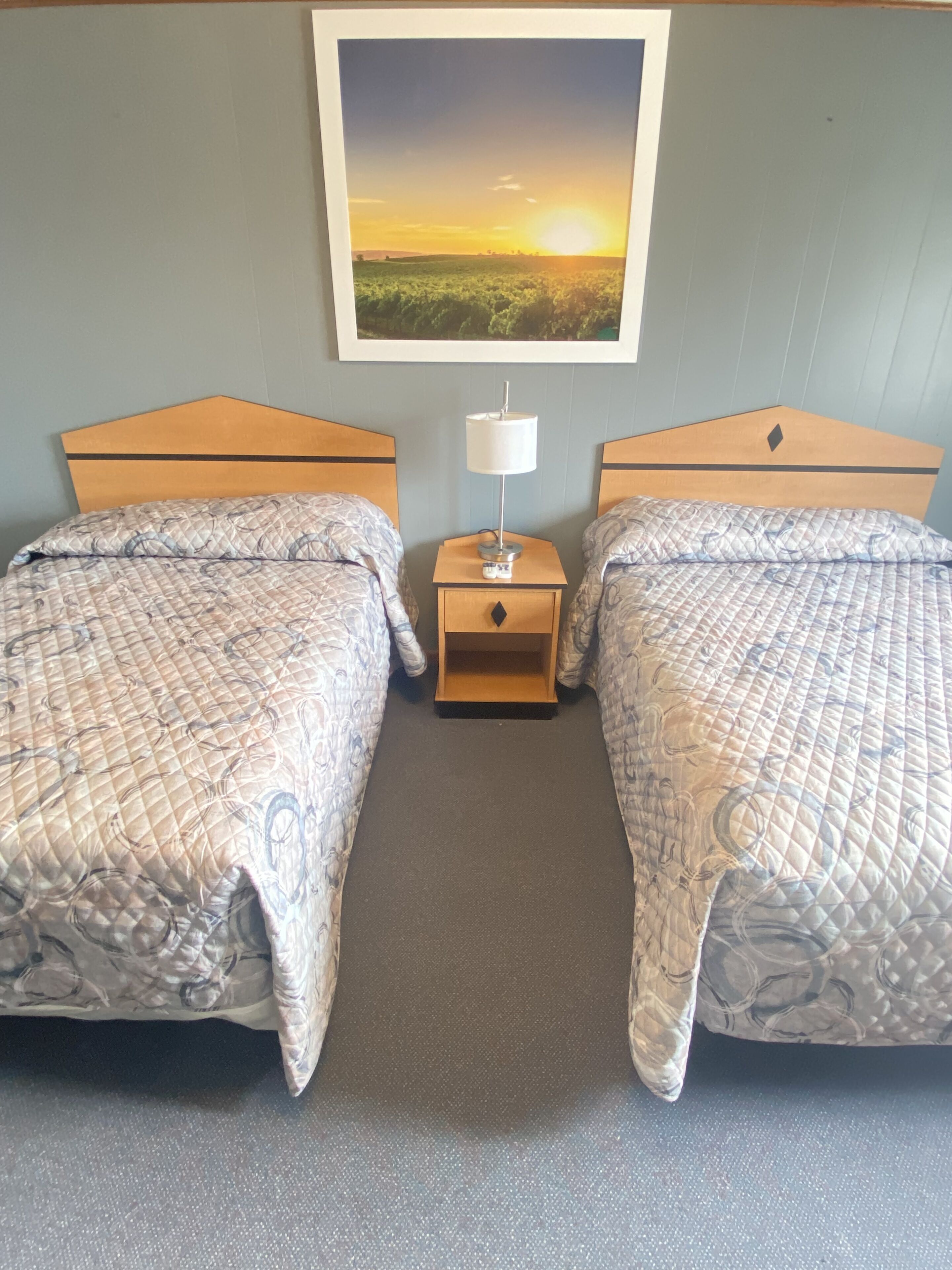 Quarto de hotel com duas camas, uma mesinha de cabeceira, uma luminária e um quadro com uma imagem de um pôr do sol sobre um campo.