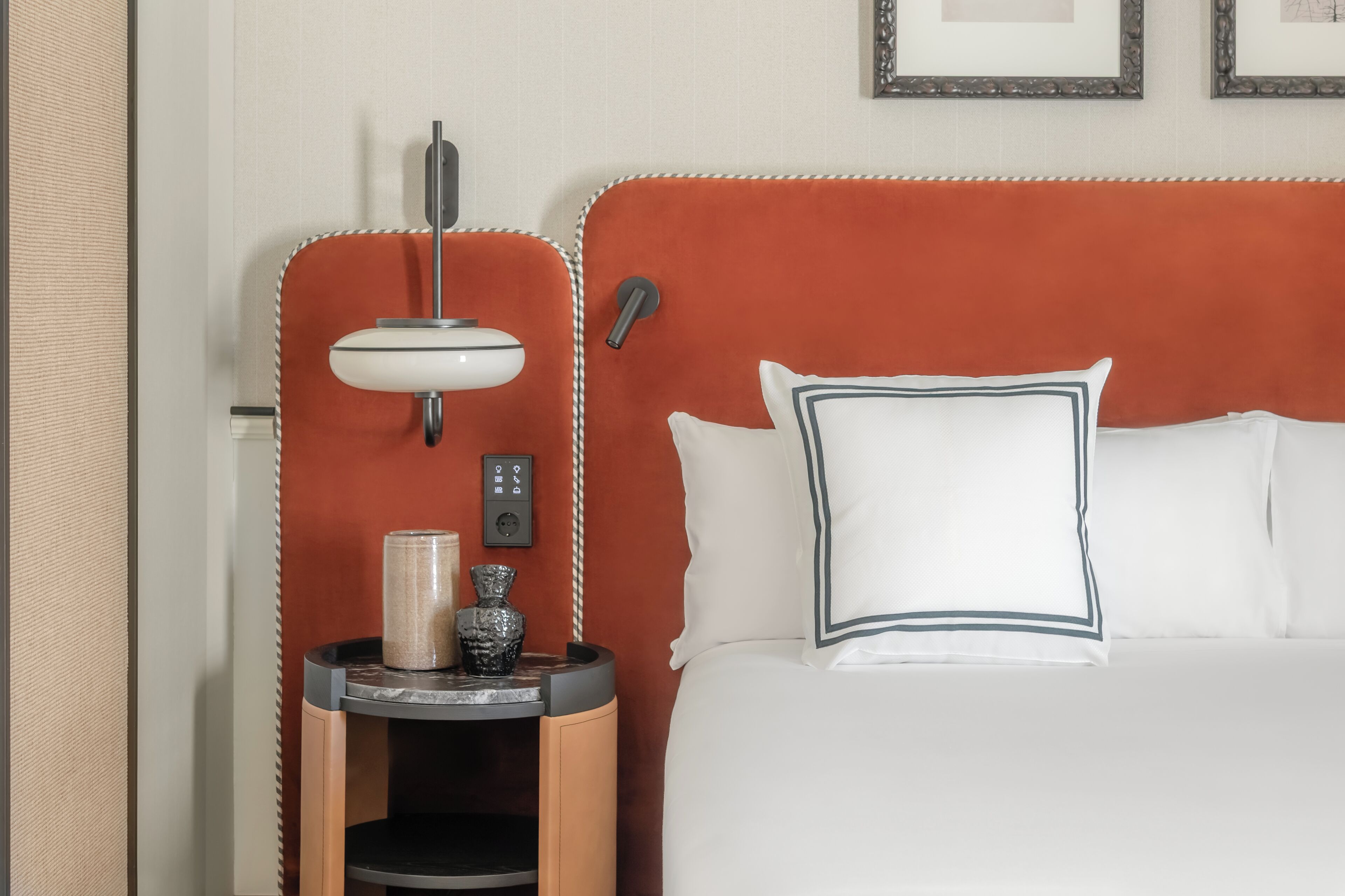A hotel room with a bed, a bedside table with a lamp, a vase, and a control panel.