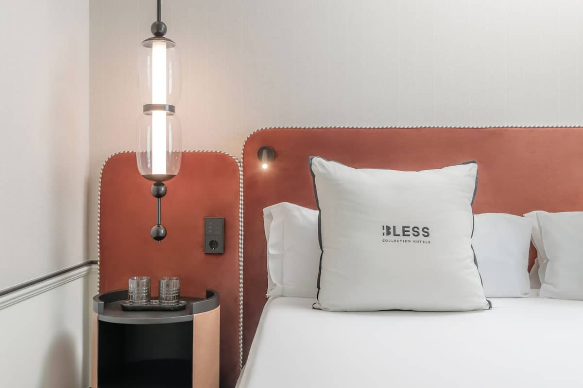 Ein ordentlich eingerichtetes Hotelzimmer mit einem Bett, einem Nachttisch mit zwei Gläsern, einer Lampe und einer Wandsteuerung.