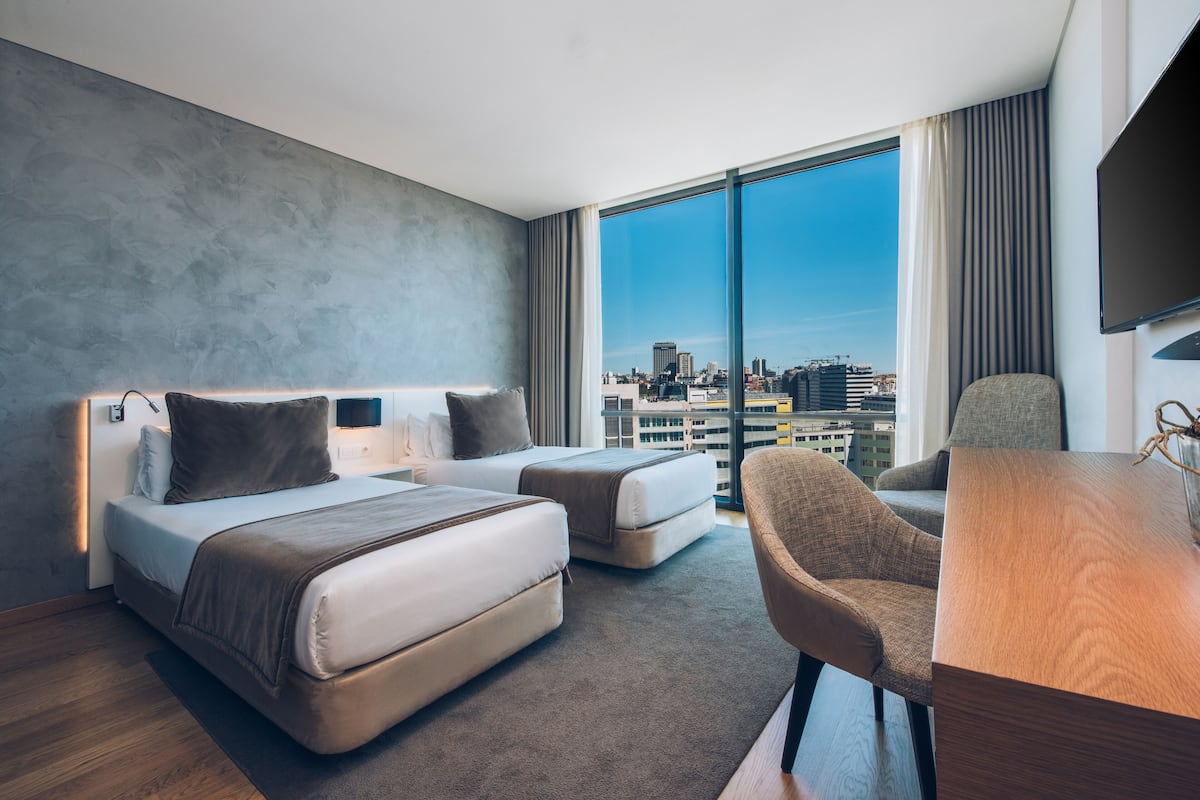 Een moderne hotelkamer met twee bedden, een bureau en een groot raam met uitzicht op de stad.