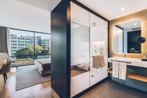 Una habitación de hotel moderna con un ventanal grande, un sofá, cabina de ducha de vidrio, tocador con espejo y una pequeña zona de estar.