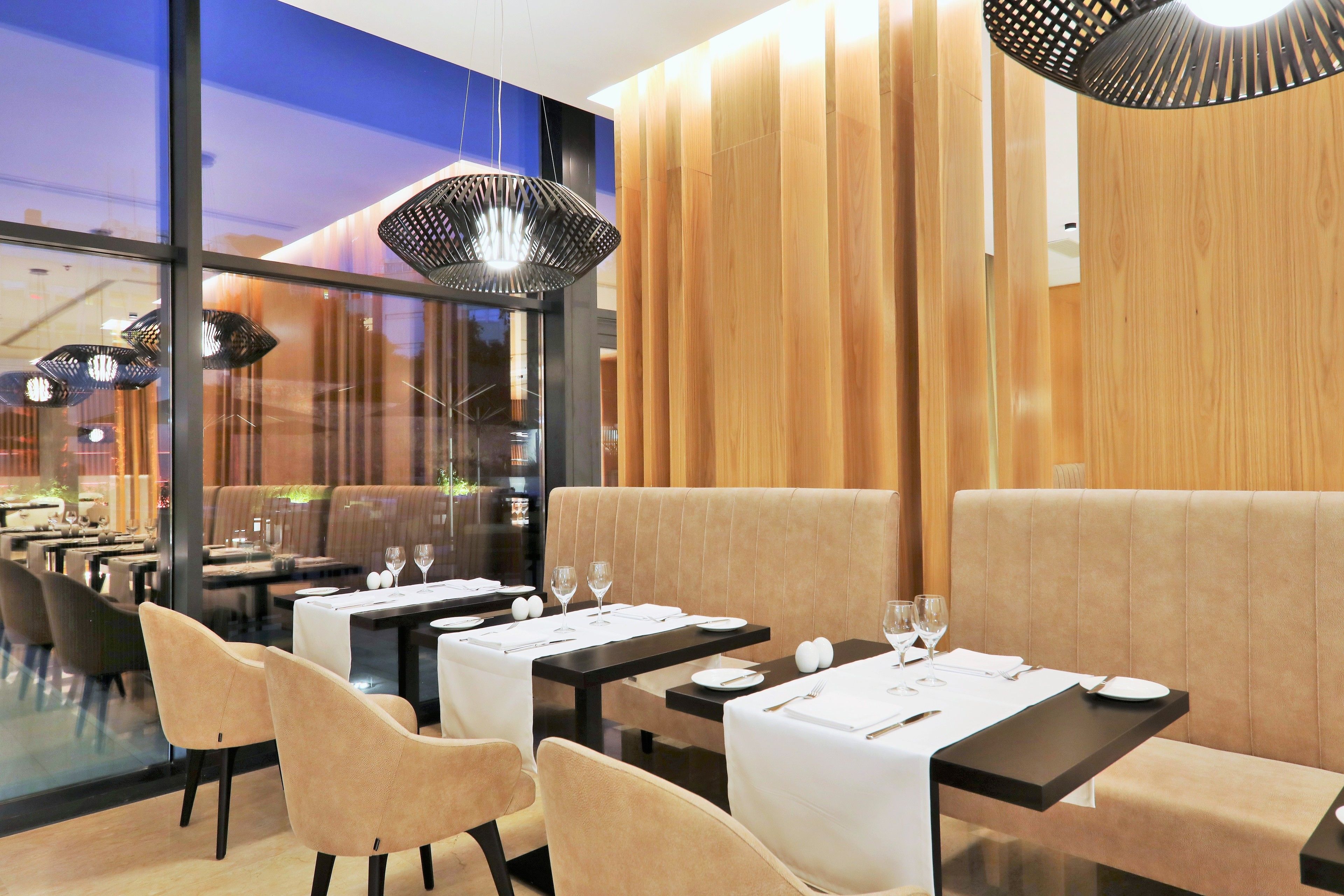 A modern restaurant interior with wooden panel walls, black tables, and beige upholstered seating.