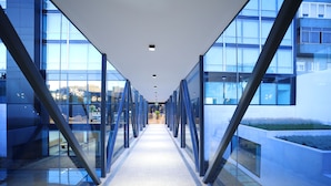 Un couloir moderne aux murs de verre et à la structure métallique, menant à l’entrée d’un bâtiment.