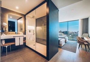 A modern hotel room with a large window offering a city view, a separate shower area, a vanity with a mirror, and a seating area with a view. - Iberostar Selection Lisboa (Lisbon)