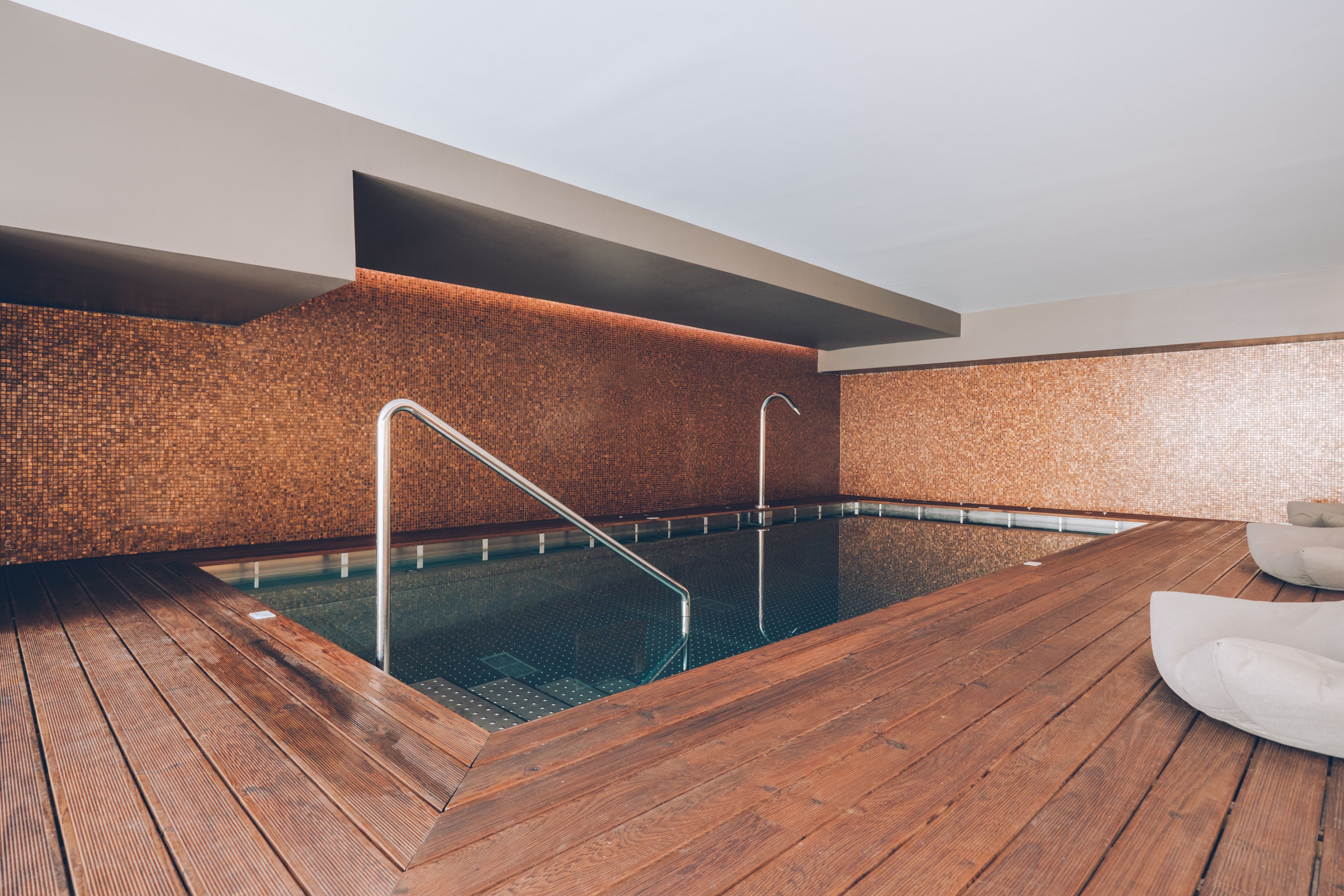 Une piscine intérieure moderne dotée d’une terrasse en bois, de rampes en métal et d’un mur à texture.