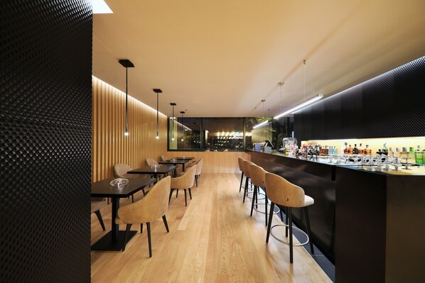 Una zona de bar moderna con una barra alargada, varios taburetes y un área de estar con mesas y sillas.