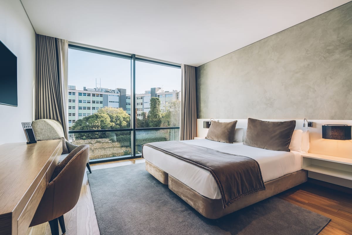 Een moderne hotelkamer met een groot bed, een bureau en uitzicht op de stad door middel van kamerhoge ramen.