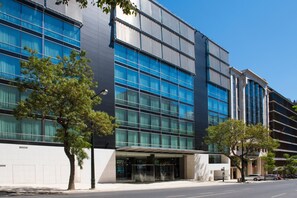 Un edificio moderno con amplios ventanales, un acceso cubierto y un árbol en la fachada.