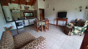 A living room with a sofa, a dining table with chairs, a television, and a kitchen area with appliances. - Cabañas Normana Inn (El Calafate)