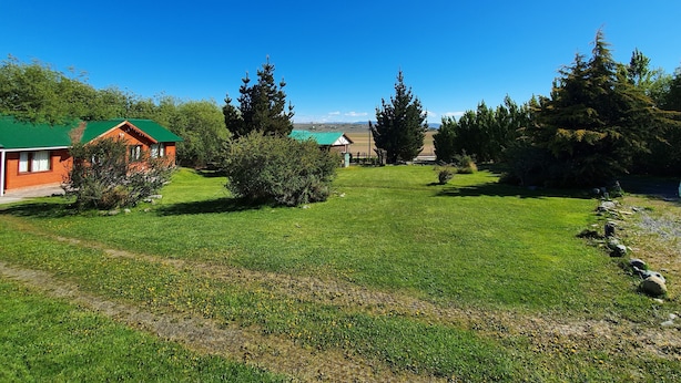 A rural landscape with a green field, a wooden cabin, and a large evergreen tree. - Cabañas Normana Inn (El Calafate)