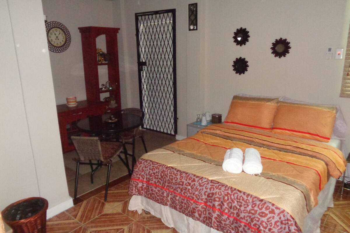 A bedroom with a bed, a desk, a chair, a small table, and a wall clock.