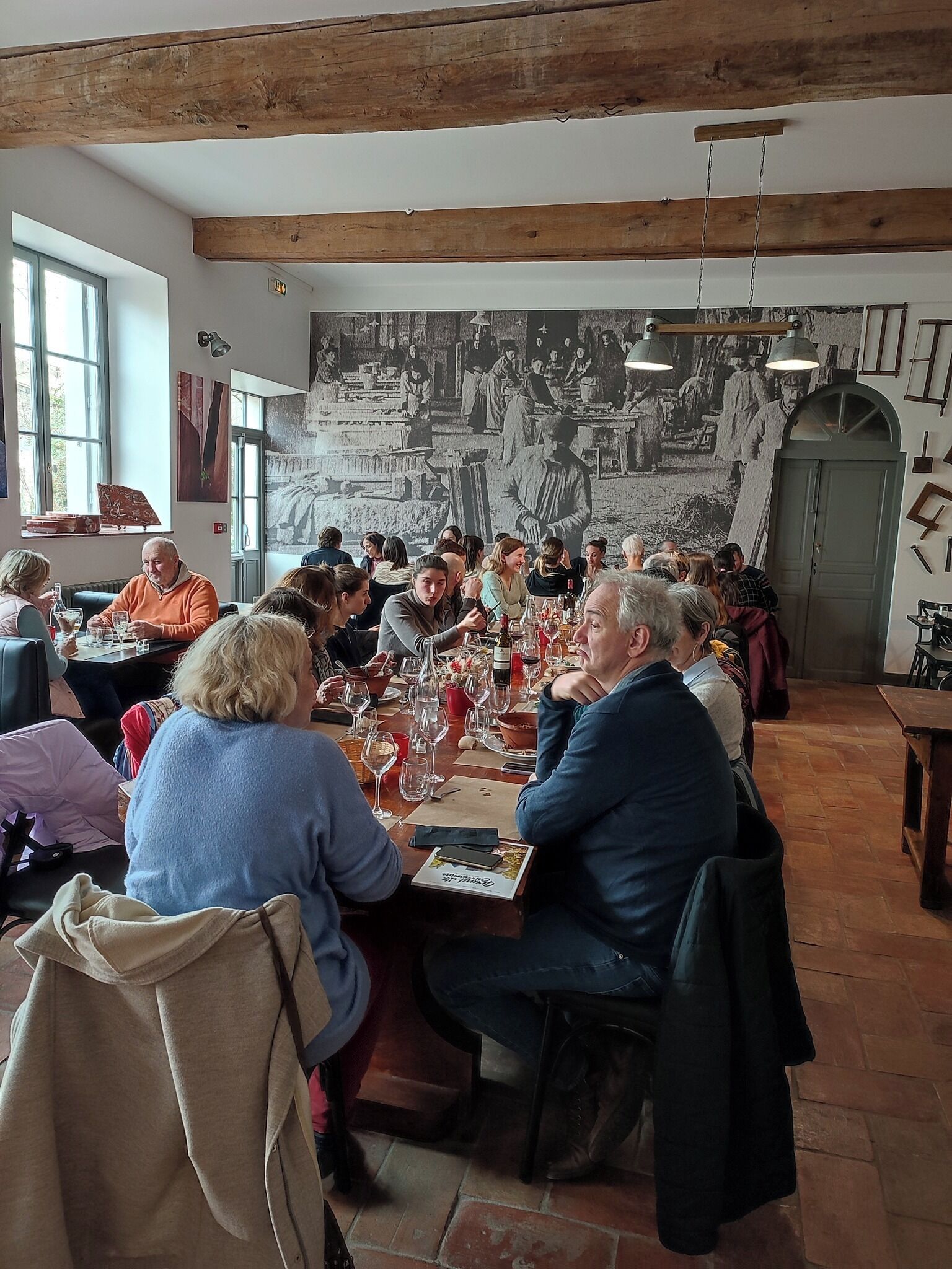 Eine Gruppe von Personen sitzt an einem Tisch in einem Raum mit freiliegenden Holzbalken, einem Wandgemälde und Blick ins Freie durch ein Fenster.
