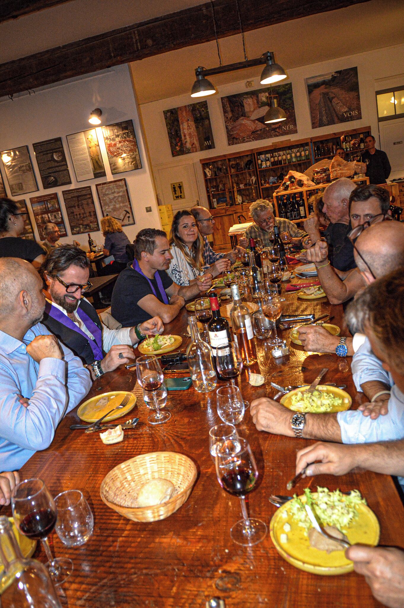 A group of people dining at a long wooden table with wine glasses and plates of food.