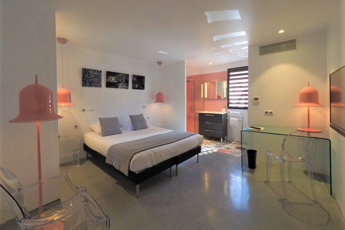 Een moderne slaapkamer met een groot bed, nachtkastjes en een glazen bureau. Er hangen twee hanglampen en een spiegel aan de muur.