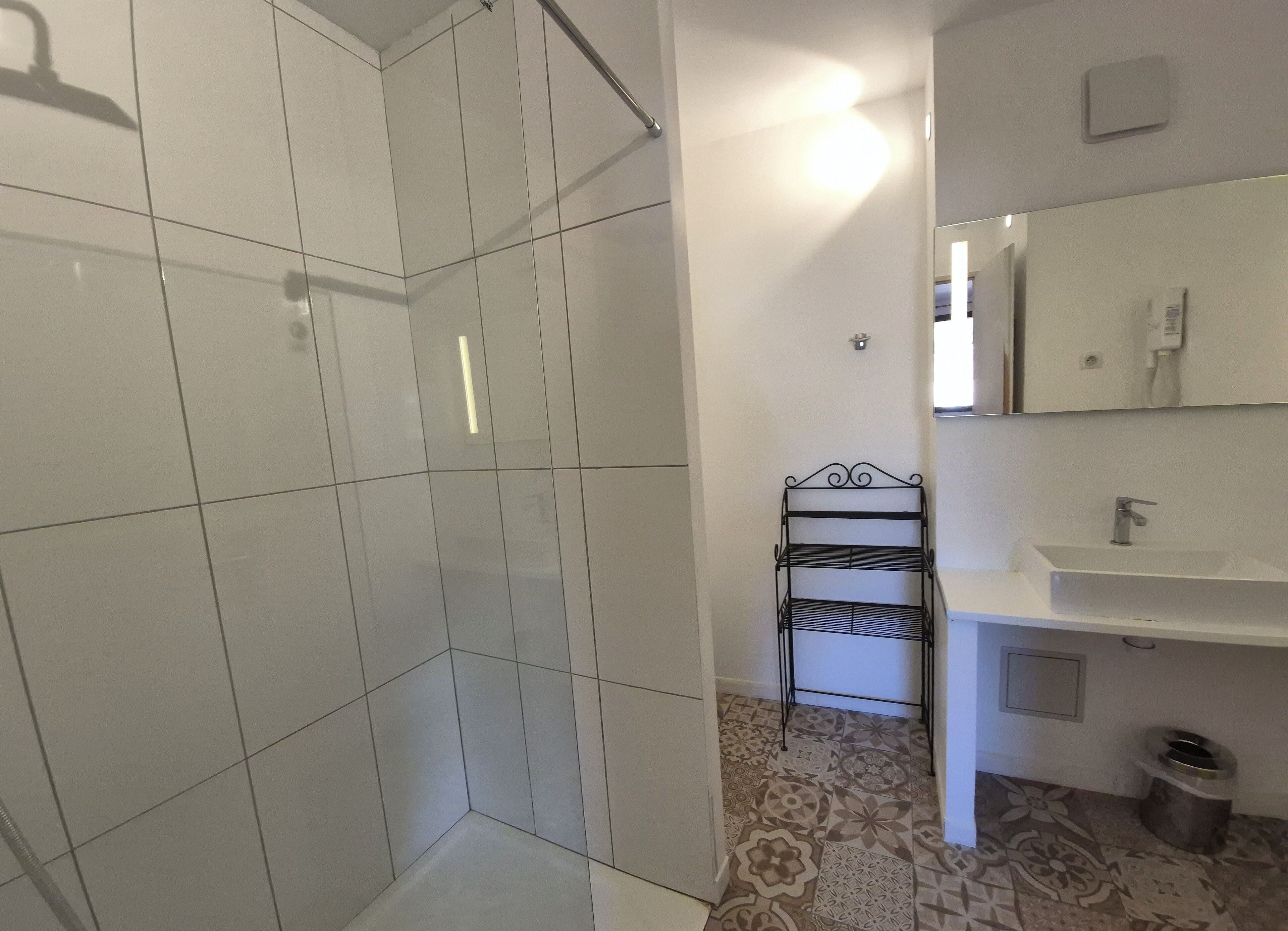 A bathroom with white tiled walls, a shower area, a sink with a mirror, and a patterned floor.