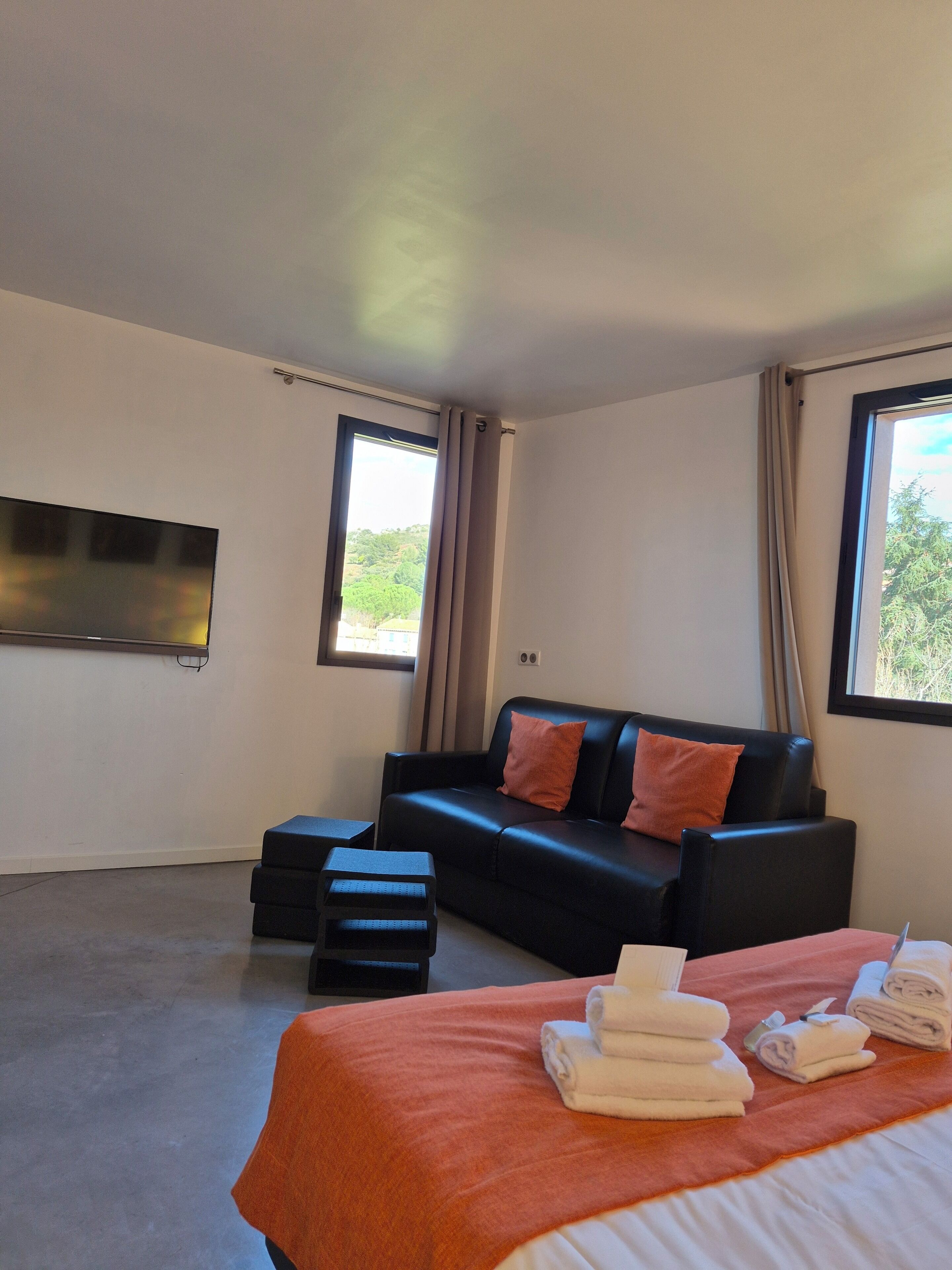 Ein modernes Hotelzimmer mit einer schwarzen Ledersofa, einem Bett mit orangefarbener Tagesdecke, einem flachbildfernseher an der Wand und einem Fenster mit Vorhängen.