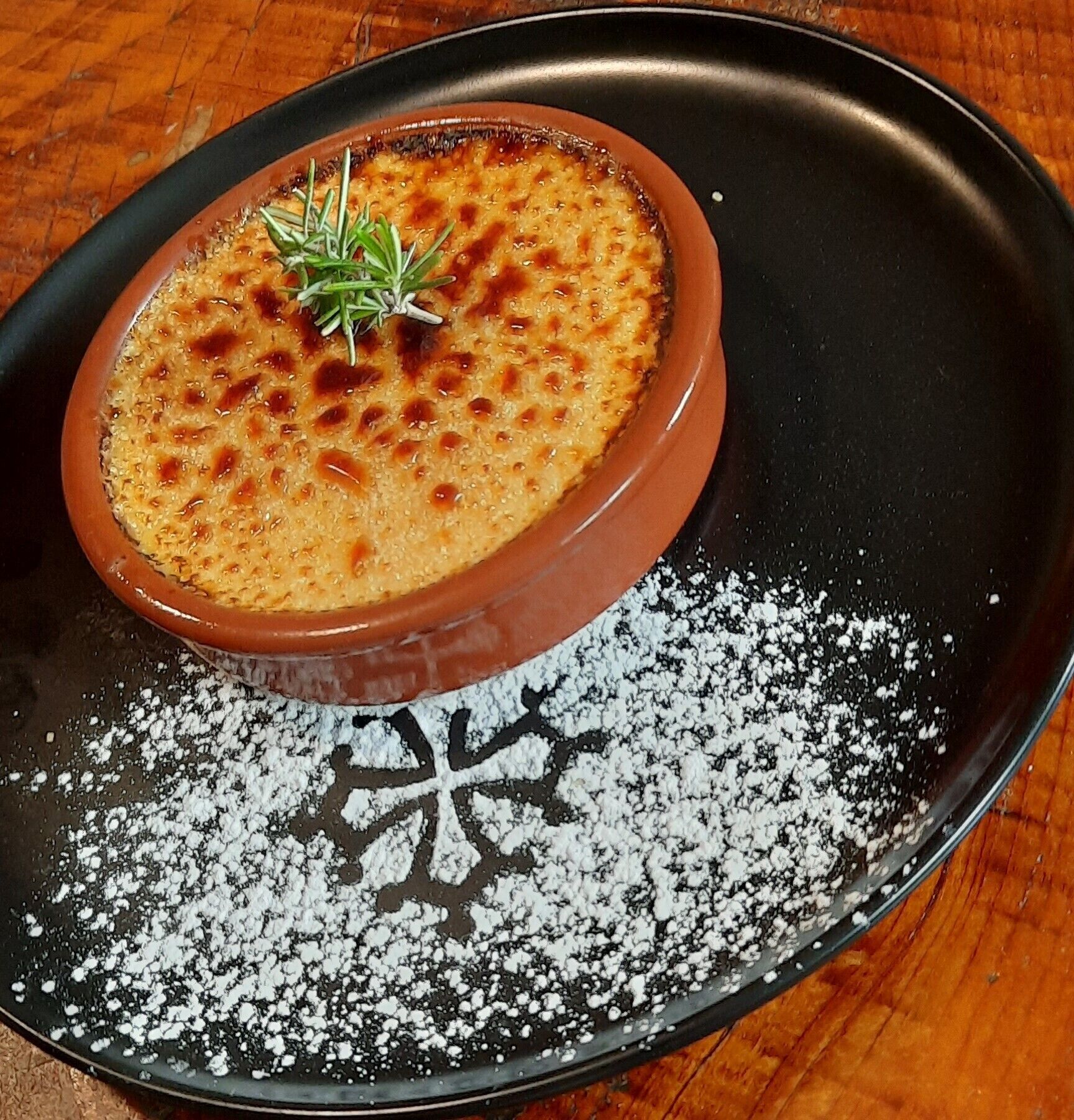 Un bol de crema brûlée sobre un plato negro con azúcar glas.
