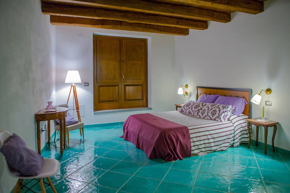 Un dormitorio con cama, dos mesitas de noche, una silla, armario de madera y ventana.