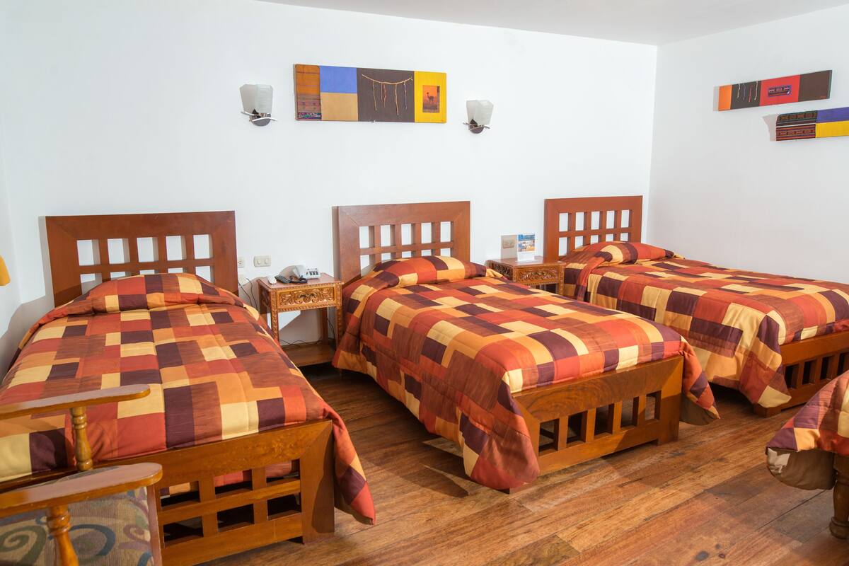 A room with three wooden beds, each with a checkered bedspread. There are wall-mounted light fixtures and a framed picture above the beds.