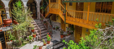 A courtyard with a staircase leading to a yellow building with a balcony. There are potted plants and a bench in the courtyard.