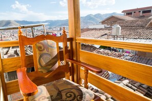 A wooden chair with a patterned cushion, a balcony railing, and a view of a cityscape with buildings and mountains in the background.