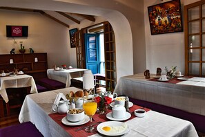 A breakfast setting with tables set for two, featuring a variety of breakfast items, a painting on the wall, and a view through an open door.
