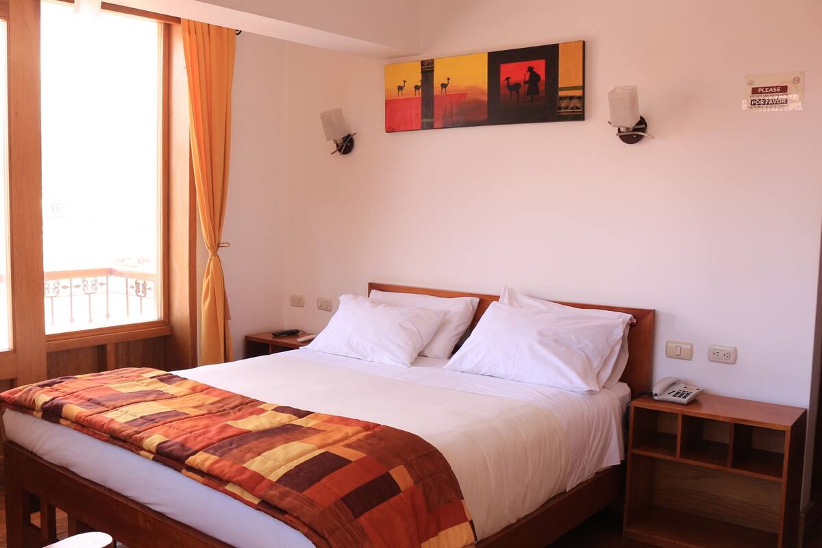 A hotel room with a bed, a nightstand, a wall-mounted lamp, a painting, and a window with curtains.
