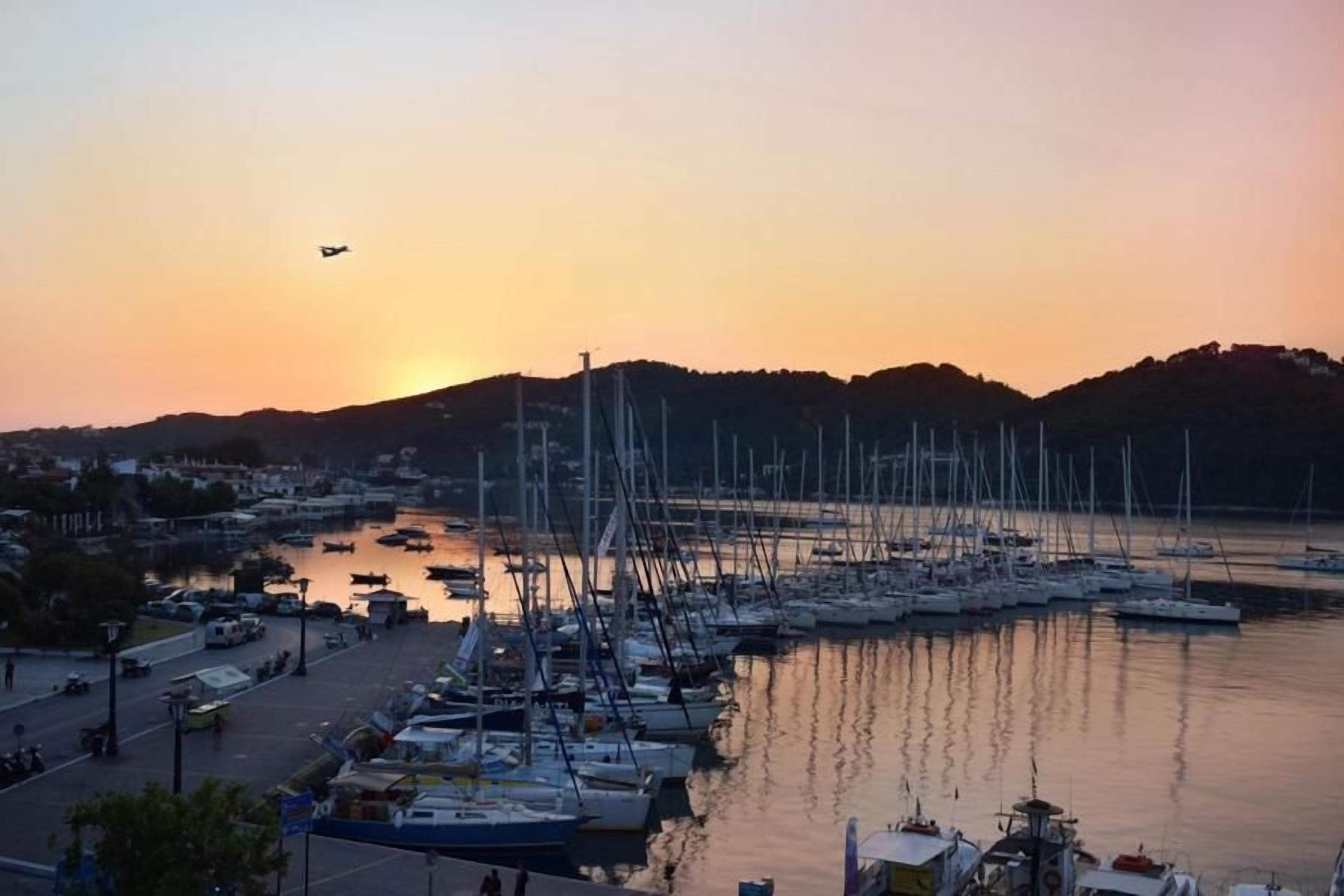 Uma marina com barcos atracados, uma estrada com veículos e um pôr do sol ao fundo.