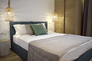 A neatly made bed with a blue headboard, white and patterned pillows, and a grey blanket. There are two bedside lamps with a grid design.