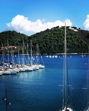 A marina with multiple sailboats docked, surrounded by a calm sea and a forested hill in the background. - Meltemi Hotel (Skiathos)