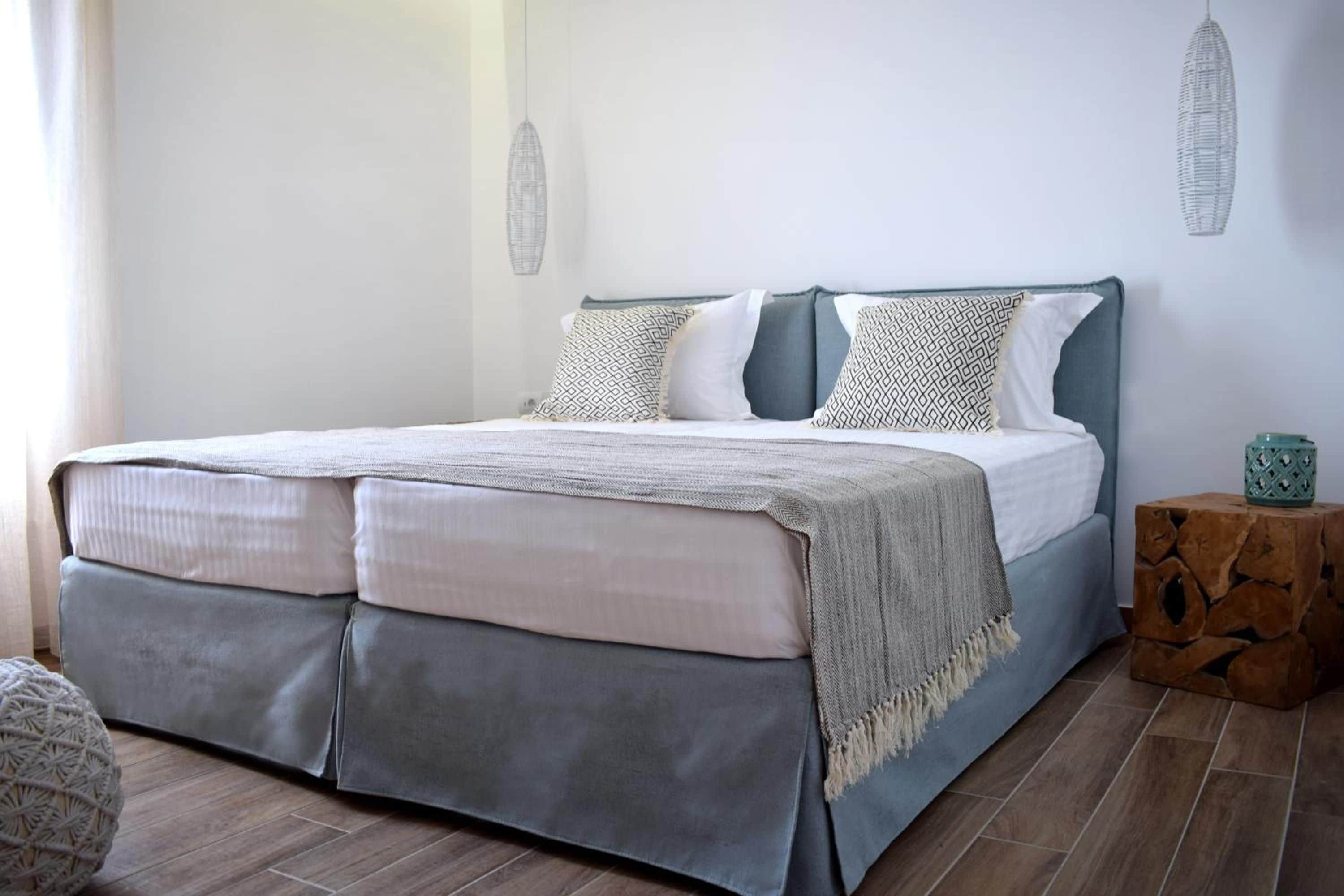 Un letto con biancheria grigio e bianca, una coperta in tessuto intrecciato e un cuscino decorativo con motivo geometrico.