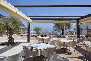 Un patio al aire libre con mesas y sillas de mimbre, un espacio de estar cubierto y vista al mar.