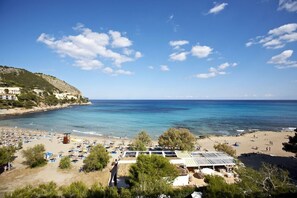 A beach with clear blue water, a sandy shore, and a few buildings in the background. - Universal Apartaments Laguna Garden (Capdepera)