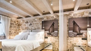 A bedroom with a stone wall, a bed, a window with curtains, and a painting of a sailboat. - Apartmani Orlando (Dubrovnik)
