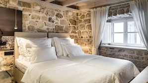 A bedroom with a stone wall, a bed with white bedding, a hanging light fixture, and a window with curtains. - Apartmani Orlando (Dubrovnik)