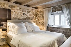 A bedroom with a stone wall, a bed with white bedding, a hanging light fixture, and a window with curtains. - Apartmani Orlando (Dubrovnik)