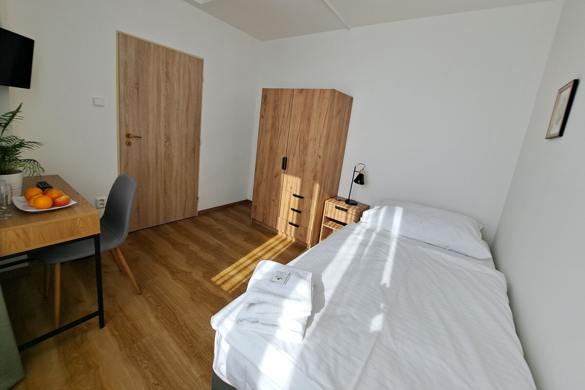 A small, well-lit room with a wooden desk, a chair, a bed with white bedding, and a large wardrobe.