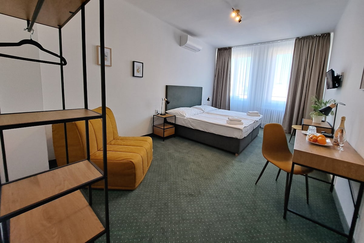 A hotel room with a bed, a desk with a chair, a sofa, a hanging chair, and a small table with a bottle and glasses.