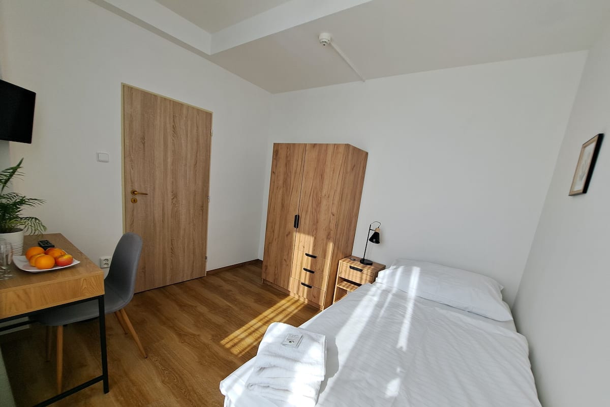 A small, well-lit room with a wooden desk, a chair, a bed with white bedding, and a large wardrobe.