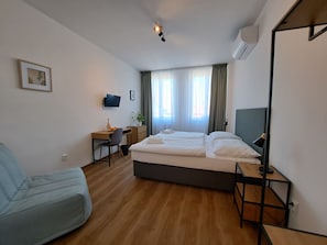 A hotel room with a bed, a desk, a chair, a TV, and a window with curtains. - Legie (Prague)