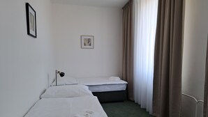 A hotel room with two single beds, a small bedside table with a lamp, a framed picture on the wall, and a window with curtains. - Legie (Prague)